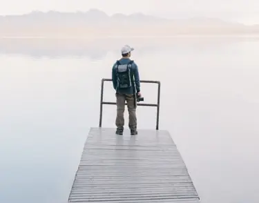 A hiker at the end of a wooden pier, facing a lake ringed by mountains.