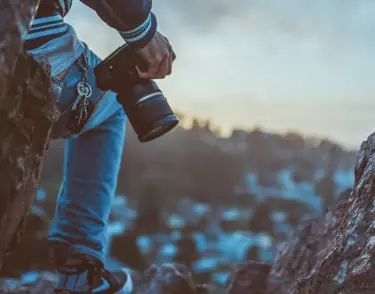 A photographer on a rock ledge at dusk, holding a DSLR at their side.