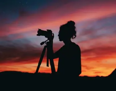 A photographer silhouetted against an orange sunset, adjusting a tripod-mounted camera.